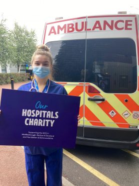 A doctor standing at the back of an ambulance holds up an Our Hospitals Charity sign