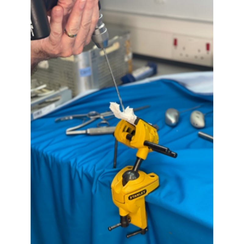 A yellow vice gripping a 3D modelled joint attached to a table with a blue cover. Someone is drilling a hole into the modelled joint.