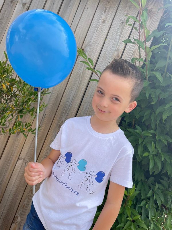 A boy holding a blue balloon models the Mackenzie Thorpe t-shirt created especially by the artist to raise funds for Our Hospitals Charity.