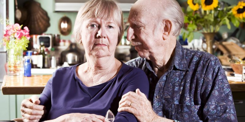 An elderly man looking at his partner with a concerned expression on his face with his arms around her comforting her.