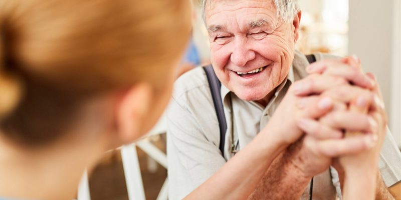 Photograph showing and elderly man smiling and a carer holding his hand, talking to him and supporting him.