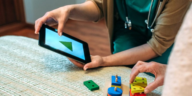 Photograph showing a health professional holding a tablet device and working alongside an elderly lady who is using shaped wooden blocks to match the shapes displayed  on the tablet screen.