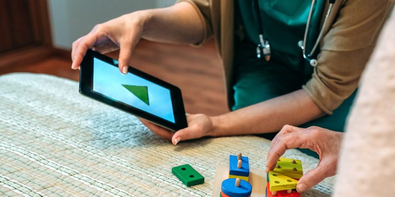 Photograph showing a health professional holding a tablet device and working alongside an elderly lady who is using shaped wooden blocks to match the shapes displayed on the tablet screen.