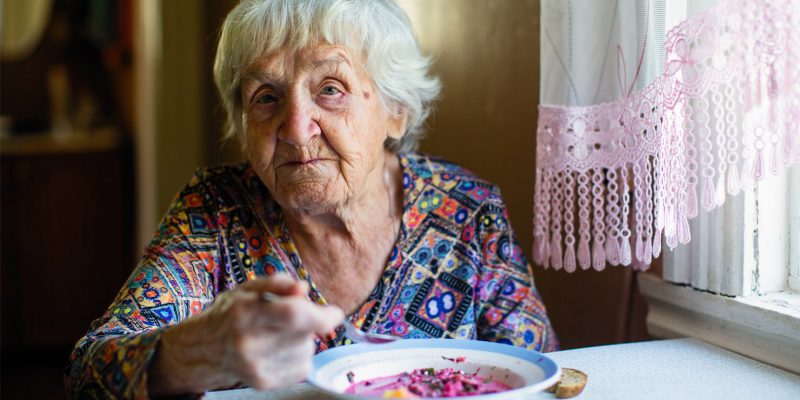 An elderly lady sat at her kitchen table eating some soup.