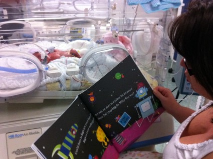 A mum reading a story book to a baby in an incubator.