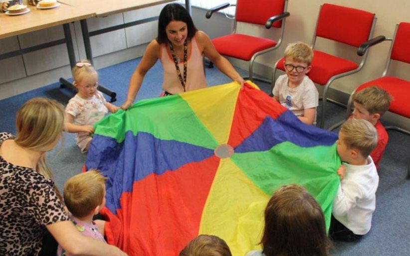Children sitting in a circle playing with a multi-coloured sheet in with speech and language therapists.