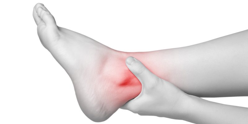 Black and white image of a person holding their ankle, the ankle area has been coloured red to signify pain.