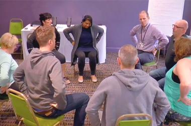 A group session with people sat on chairs in a circle - all are holding their lower back.