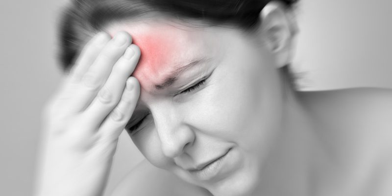 A black and white image of someone with a painful expression on their face holding their forehead. The forehead has been coloured ref to signify pain.