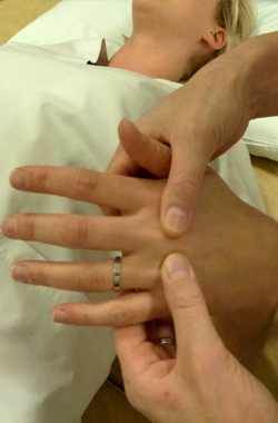 The hand is palm facing toward side of the body - the physiotherapist is applying pressure with both thumbs across the top of the hand, just above the knuckles