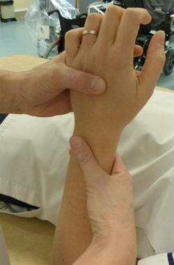 Hand is being held with the palm facing toward the body, the physiotherapist is applying pressure with their thumb to the top of the hand. The wrist is being supported with the physiotherapists other hand