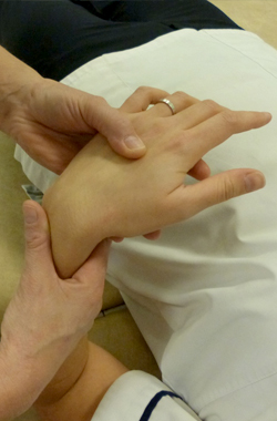 closeup of the hand. Wrist is supported and the hand is palm toward stomach - the physiotherapist is applying pressure with their thumb across the top of the hand, just above the knuckles