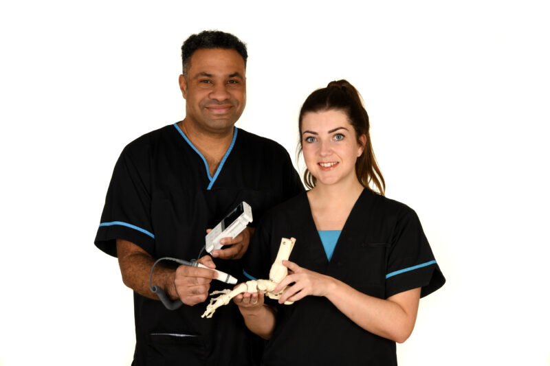 Two podiatrists. One holds podiatry equipment, scanning an artificial skeleton of a foot, held by the other