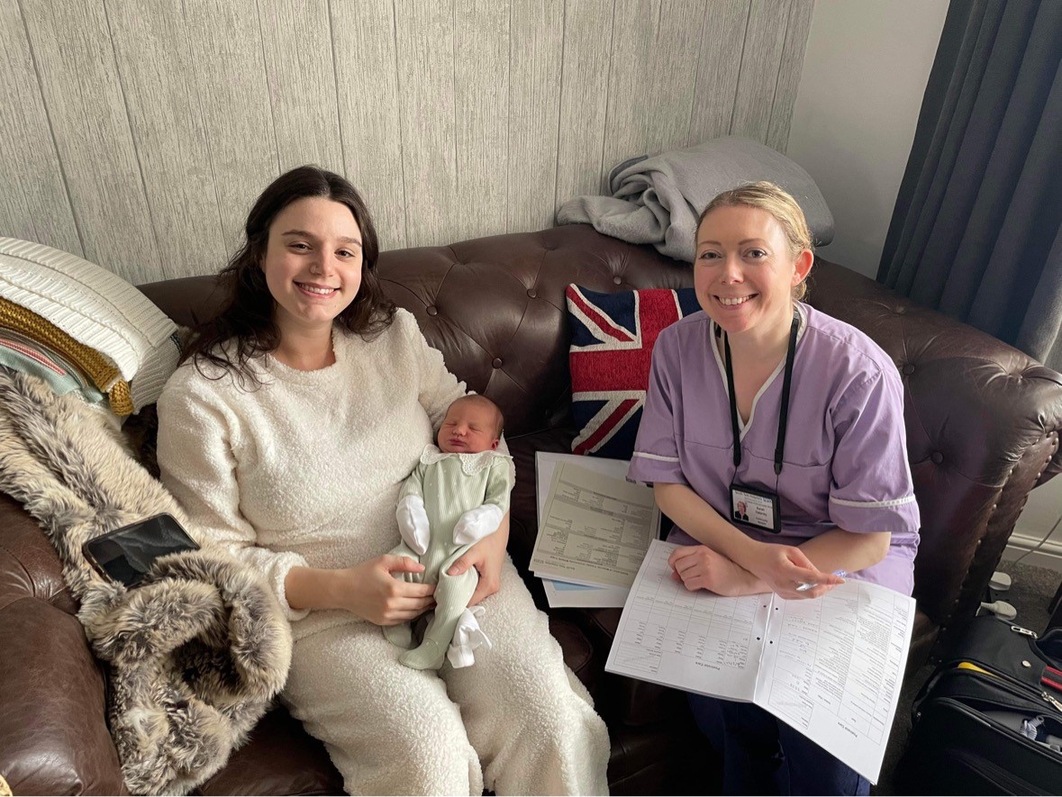 Image of community midwife smiling, sitting next to a new parent who is also smiling whilst cradling their baby