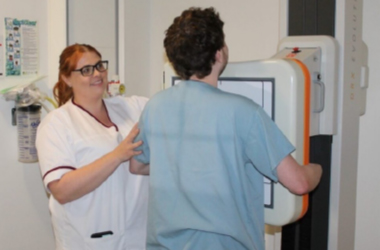 A radiographer with their patient who is stood against the machine preparing to have an x-ray